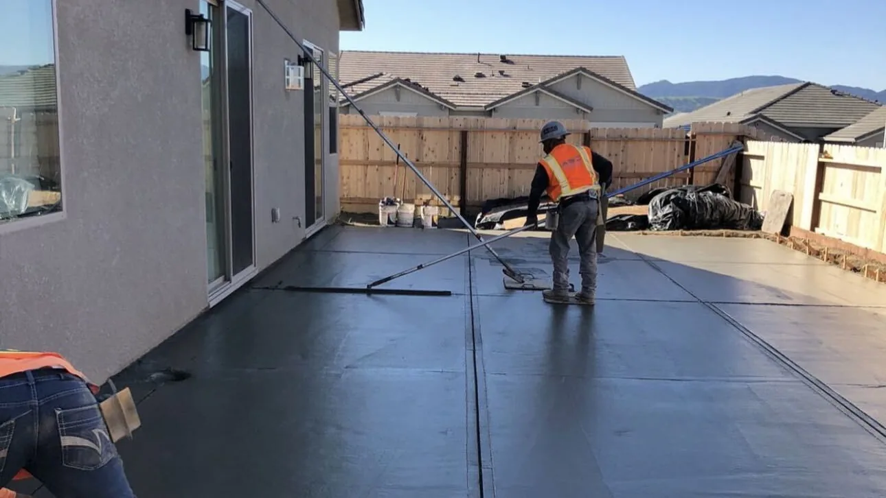 Two-man crew bull-floating a wet concrete slab along the back of a house