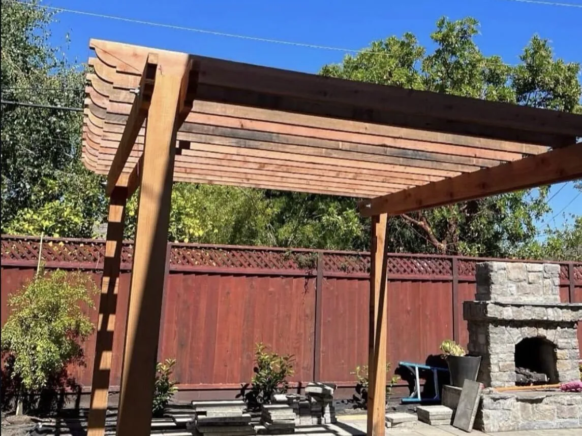 Redwood pergola beside a stone outdoor fireplace in a fenced backyard